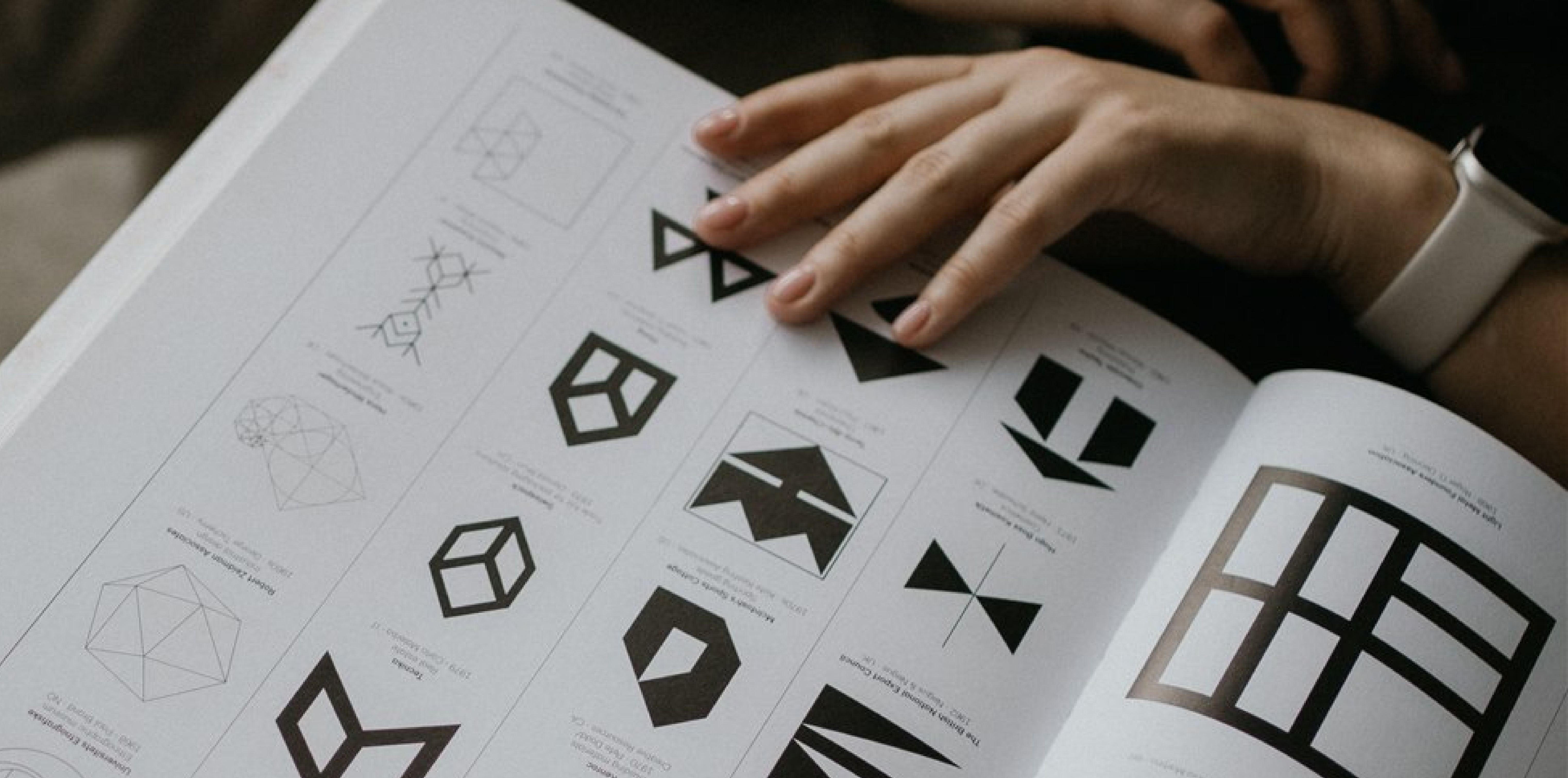 A close-up of a design book featuring various geometric logo concepts and shapes, with a hand turning the pages, showcasing creativity and design exploration.