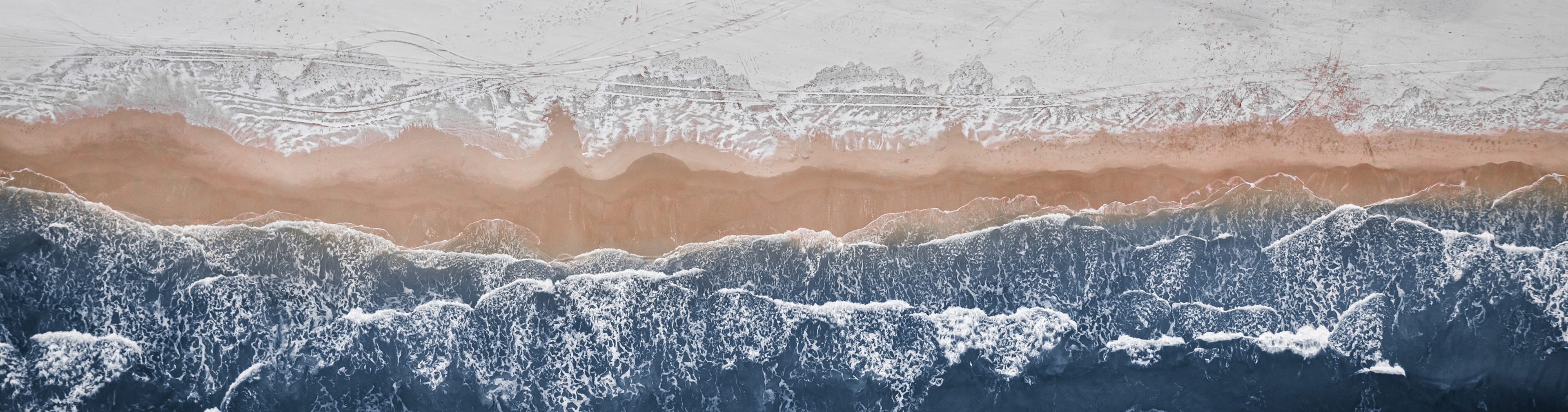 Aerial view of winter shoreline with waves crashing on a snowy beach, combining textures of ocean, sand, and snow for a dramatic natural landscape.