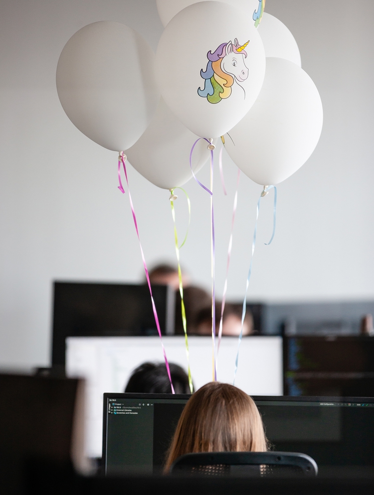 Office scene with a female developer working at a computer, surrounded by unicorn balloons, symbolizing creativity and team culture in tech.